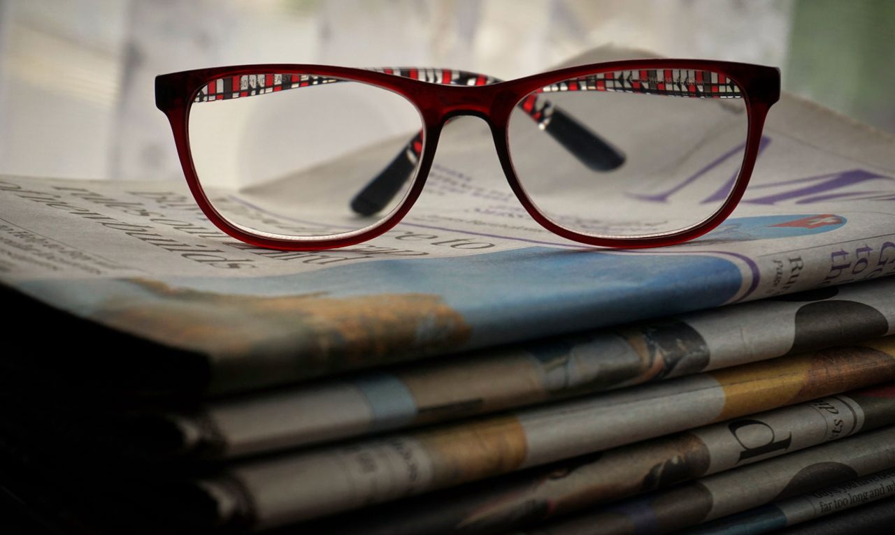 Close-up of red glasses resting on stacked newspapers, emphasizing reading and vision.
